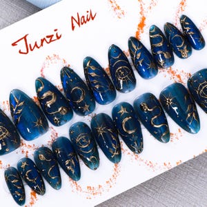 Puede incluir: Un conjunto de uñas postizas azules y doradas con diseños celestiales. Cada uña presenta una base azul oscuro con detalles dorados, incluyendo lunas, estrellas y patrones abstractos. El texto "Junzi Nail" está escrito en rojo.
