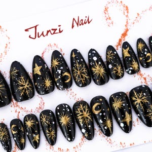Puede incluir: Uñas postizas negras con diseños celestiales dorados, que incluyen estrellas, lunas y copos de nieve. Las uñas tienen forma de almendra y presentan un acabado brillante. El texto "Junzi Nail" está escrito en escritura roja.