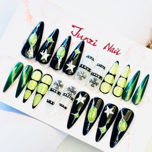 Puede incluir: Un conjunto de 12 uñas postizas negras y verdes con una variedad de diseños, incluyendo estrellas, cruces y círculos verdes. Las uñas están dispuestas sobre una superficie blanca con el texto "Junzi Nail" en rojo.