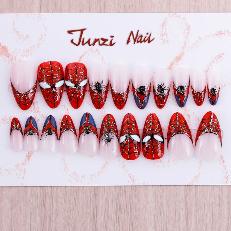 Puede incluir: Un conjunto de 20 u&ntilde;as postizas con tem&aacute;tica de Spiderman. Las u&ntilde;as son de forma ovalada y tienen un dise&ntilde;o de telara&ntilde;a roja y negra con una ara&ntilde;a y los ojos de Spiderman. Las u&ntilde;as se muestran en una tarjeta blanca con el texto "Junzi Nail".