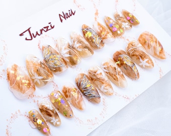 May include: A set of 14 long, almond-shaped false nails with a brown, translucent base. Each nail is decorated with gold glitter, white lines, and a silver leaf design. The nails are arranged on a white surface with the text "Junzi Nail" in red.