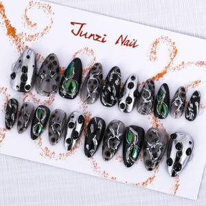 May include: A set of 14 false nails with a black and silver marble design. The nails have black and silver accents and some have a green iridescent accent. The nails are displayed on a white card with the text "Junzi Nail".