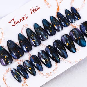 Puede incluir: Un conjunto de 10 uñas postizas con un diseño de galaxia. Las uñas son negras con remolinos azules, morados y blancos, y tienen purpurina dorada y estrellas. Las uñas están dispuestas en una tarjeta blanca con el texto "Junzi Nail" en rojo.