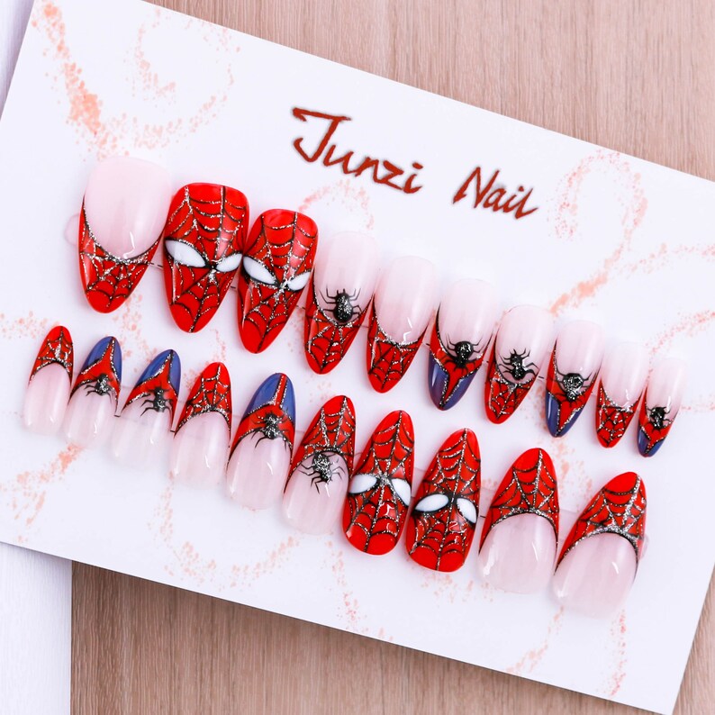 Puede incluir: Un conjunto de 12 u&ntilde;as postizas con un dise&ntilde;o de telara&ntilde;a roja y negra. Las u&ntilde;as son de forma ovalada y tienen un acabado brillante. El dise&ntilde;o presenta una telara&ntilde;a con una ara&ntilde;a en el centro. El texto "Junzi Nail" est&aacute; impreso en la tarjeta blanca.