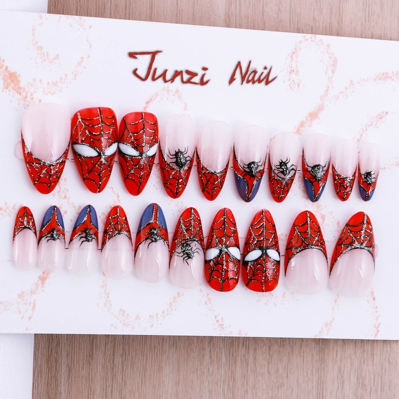 Puede incluir: Conjunto de 20 u&ntilde;as postizas con un dise&ntilde;o de Spiderman. Las u&ntilde;as son de forma ovalada y tienen un dise&ntilde;o de telara&ntilde;a roja y negra con un ojo de ara&ntilde;a blanco. Las u&ntilde;as est&aacute;n dispuestas en dos filas en una tarjeta blanca con el texto "Junzi Nail" impreso en ella.