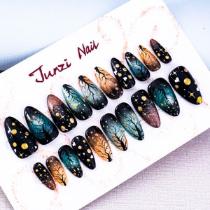 Puede incluir: Un conjunto de 12 uñas postizas con un diseño degradado en azul oscuro, verde y dorado. Las uñas presentan una silueta de árbol caprichosa y una dispersión de estrellas doradas. Las uñas se muestran en una tarjeta blanca con el texto "Junzi Nail" en rojo.