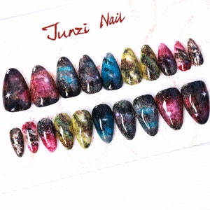 Puede incluir: Un conjunto de 18 uñas postizas con un diseño de galaxia. Las uñas son de varios tonos de azul, morado, rosa y dorado con detalles de purpurina. Las uñas están dispuestas en dos filas sobre una superficie blanca. El texto "Junzi Nail" está escrito en rojo en la parte superior de la imagen.