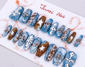 Puede incluir: Un conjunto de 10 uñas postizas azules, marrones y blancas con un diseño de mezclilla, leopardo y estrella. Las uñas presentan abalorios de cruz plateados y pedrería. Junzi Nail está escrito en la tarjeta blanca.