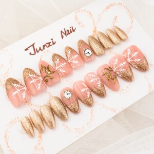 May include: A set of 10 false nails with a pink and gold design. The nails feature a snowflake design with gold glitter and a clock face accent. The nails are displayed on a white card with the text "Junzi Nail" written in black.