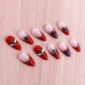 Puede incluir: Diez puntas de u&ntilde;as rojas y blancas con un dise&ntilde;o de telara&ntilde;a negra. Las puntas de u&ntilde;as est&aacute;n dispuestas en una fila sobre una superficie de madera marr&oacute;n claro.