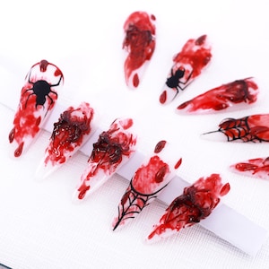 Può includere: Unghie finte a tema Halloween. Le unghie bianche presentano dettagli rossi e neri, con disegni di ragni, ragnatele e schizzi di sangue. Disposte su una superficie bianca.