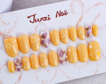 May include: A set of 12 yellow press-on nails with white and purple floral designs. The nails are arranged in two rows on a white card with the text "Junzi Nail" in red.