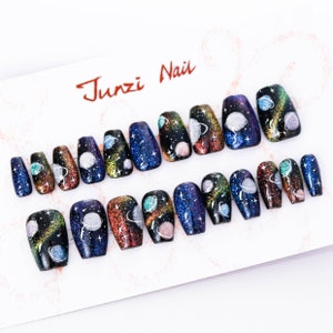 Puede incluir: Un conjunto de 18 uñas postizas con temática de galaxia. Las uñas están decoradas con purpurina y pintadas con un fondo azul oscuro, negro y morado con planetas y estrellas plateados y dorados.
