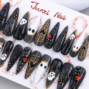 Può includere: Un set di unghie finte lunghe e appuntite a tema Halloween. Le unghie sono nere, grigie e bianche, con accenti dorati. I disegni includono pipistrelli, ragni, ragnatele, fantasmi e zucche. Il testo "Junzi Nail" è visibile.