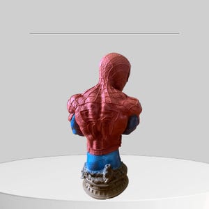 Puede incluir: Un busto de pl&aacute;stico rojo y azul de Spider-Man. El busto est&aacute; posado con la espalda del personaje hacia el espectador. El busto est&aacute; sobre una base marr&oacute;n.