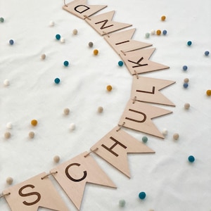 Könnte beinhalten: Holzbanner mit dem Wort "SCHULKIND" auf einzelnen Flaggen. Die Flaggen sind mit einer Schnur verbunden und mit kleinen, bunten Pompons verziert.