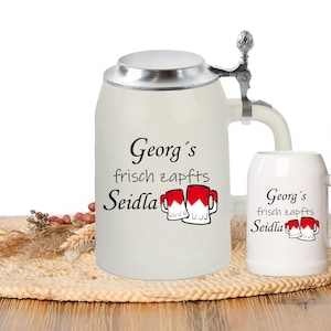 Könnte beinhalten: Ein weißer Keramik-Bierkrug mit silbernem Deckel und Henkel. Der Krug trägt den Schriftzug "Georg's frisch zapfts Seidla" und zwei rot-weiße Bierkrüge sind darauf gedruckt. Ein kleinerer weißer Keramik-Bierkrug mit dem gleichen Schriftzug und zwei Bierkrügen ist ebenfalls abgebildet.