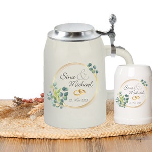 Könnte beinhalten: Cremefarbenes Keramik-Bierkrug- und Becher-Set. Der Krug hat einen Klappdeckel und einen Henkel. Beide zeigen ein goldgerahmtes Design mit den Namen "Sina & Michael", Eheringen und dem Datum "22. Mai 2023", umgeben von grünen Blättern.