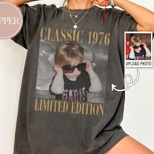 May include: Dark gray t-shirt with a photo of a child wearing sunglasses. The shirt has the text "CLASSIC 1976 LIMITED EDITION" in gold lettering. A small photo with the text "UPLOAD PHOTO" is on the right side of the shirt.