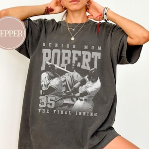 May include: Dark gray t-shirt with a baseball-themed graphic. The graphic includes the text "SENIOR MOM ROBERT 35 THE FINAL INNING" above a black and white image of baseball players.