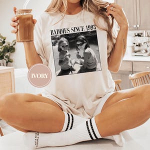 K&ouml;nnte beinhalten: Ein wei&szlig;es T-Shirt mit dem Text "Baddies Since 1993" und einem Schwarzwei&szlig;foto von zwei M&auml;dchen. Das Shirt ist in Elfenbein erh&auml;ltlich.