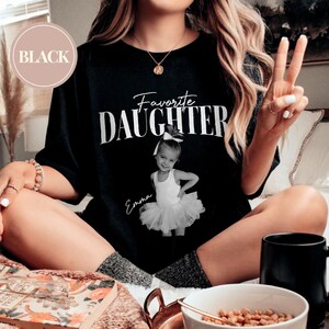 May include: Black t-shirt with the words "Favorite Daughter" in white script. A black and white photo of a young girl in a tutu is printed below the text, with the name "Emma" in script. The shirt is worn by a person.