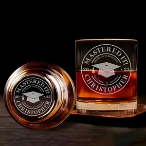 May include: Two clear glass tumblers with a brown liquid inside. Both glasses have an etched design that says "Mastered It!" with a graduation cap and the text "Bachelor of Biography 2025 University of Michigan Christopher".