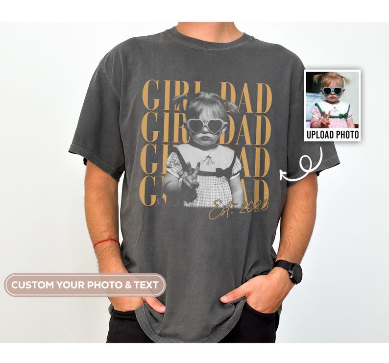 May include: Dark gray t-shirt with a graphic of a child wearing heart-shaped sunglasses and the words "GIRL DAD" in gold. The shirt also features the text "Est. 2020". A small photo is on the right side of the shirt.