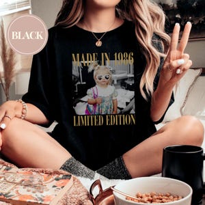 May include: Black t-shirt with a gold graphic that says "MADE IN 1986 LIMITED EDITION." The graphic is a black and white photo of a young girl wearing heart-shaped sunglasses. The shirt is worn by a person sitting down.