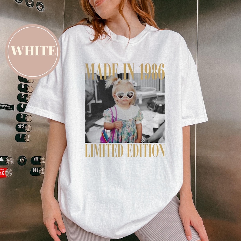 May include: White t-shirt with a black and white photo of a young child wearing heart-shaped sunglasses. The text "MADE IN 1986" and "LIMITED EDITION" are printed in gold. The shirt is a casual, oversized style.