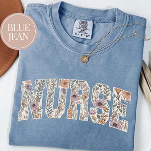 May include: A light blue t-shirt with the word "NURSE" in large, floral-filled letters. The shirt has a crew neck and short sleeves. A gold necklace with a pendant is draped across the shirt. The shirt is labeled "BLUE JEAN".