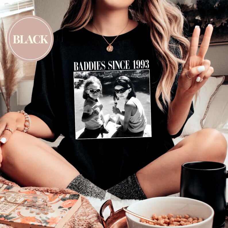 K&ouml;nnte beinhalten: Schwarzes T-Shirt mit dem Text "Baddies Since 1993" und einem Schwarzwei&szlig;foto von zwei M&auml;dchen mit Sonnenbrillen.