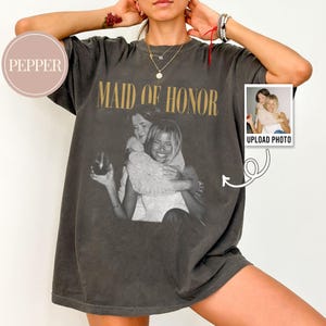 K&ouml;nnte beinhalten: Ein anthrazitfarbenes Oversize-T-Shirt mit einem schwarz-wei&szlig;en Vintage-Foto von zwei sich umarmenden Personen und dem Text "MAID OF HONOR" in gro&szlig;en goldenen Serifenschrift-Buchstaben dar&uuml;ber. Das T-Shirt enth&auml;lt ein kleines kreisf&ouml;rmiges "PEPPER"-Logo.