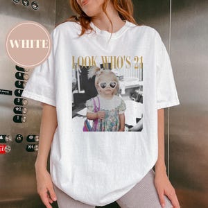 May include: White t-shirt with a graphic featuring a black and white photo of a young girl wearing heart-shaped sunglasses. The text "LOOK WHO'S 21" is printed above the photo in gold lettering.