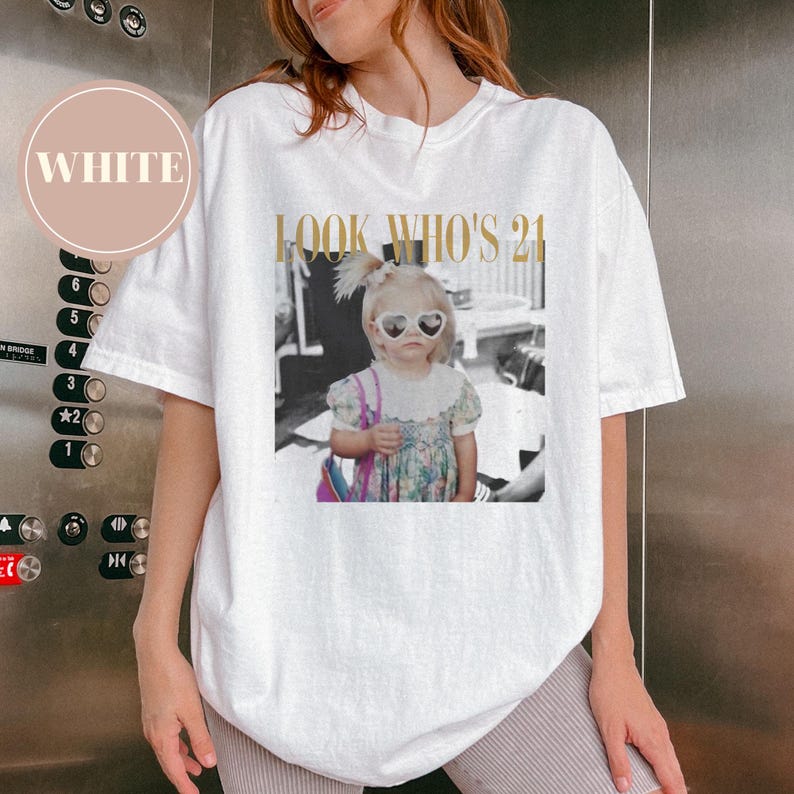 May include: White t-shirt with a graphic featuring a black and white photo of a young girl wearing heart-shaped sunglasses. The text "LOOK WHO'S 21" is printed above the photo in gold lettering.