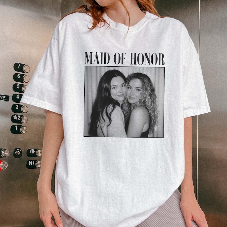 Op de afbeelding: Wit T-shirt met een zwart-wit foto van twee vrouwen en de tekst "MAID OF HONOR" erboven. De vrouwen lachen en omarmen elkaar. Het shirt heeft een casual stijl.