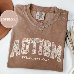 May include: A brown t-shirt with the word "AUTISM" in large, white letters outlined in black. The letters are filled with a floral pattern. Below the word "AUTISM" is the word "mama" in a smaller, white font. The shirt is made of a soft, comfortable material.