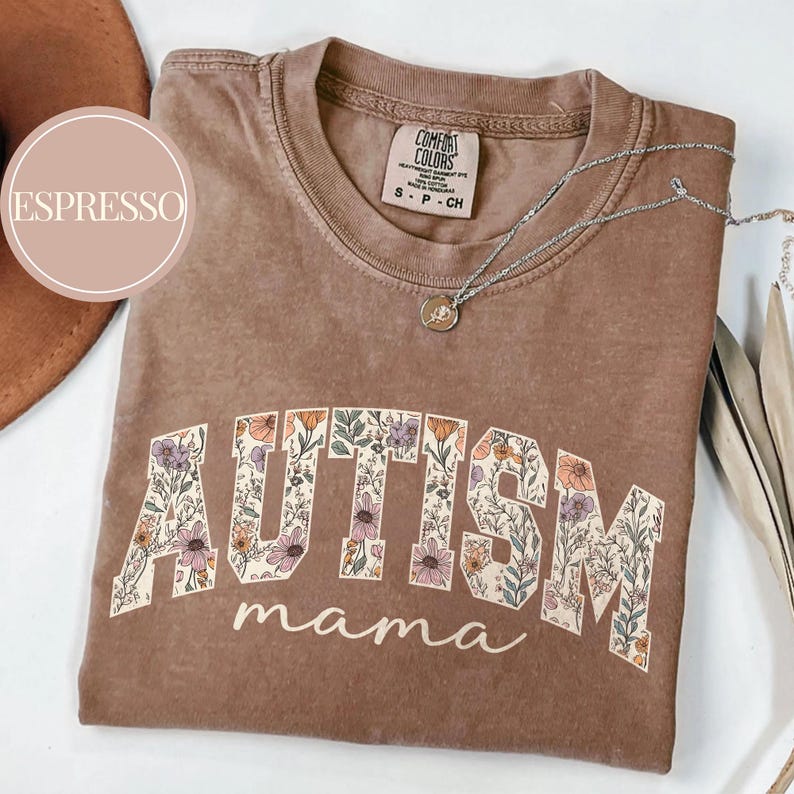 May include: A brown t-shirt with the word "AUTISM" in large, white letters outlined in black. The letters are filled with a floral pattern. Below the word "AUTISM" is the word "mama" in a smaller, white font. The shirt is made of a soft, comfortable material.