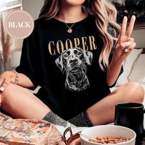 May include: Black t-shirt featuring a detailed illustration of a dog's face with the name "COOPER" in gold lettering above. A circular "BLACK" label is in the upper left corner. The shirt is a casual style.