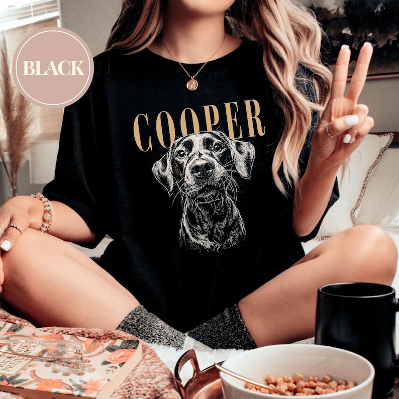 May include: Black t-shirt featuring a detailed illustration of a dog's face with the name "COOPER" in gold lettering above. A circular "BLACK" label is in the upper left corner. The shirt is a casual style.
