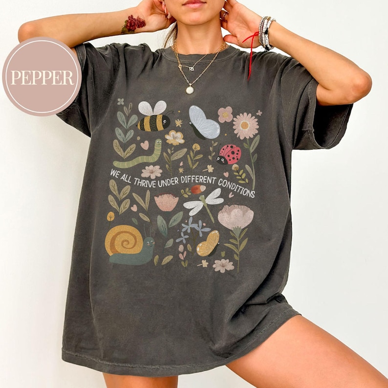 May include: Dark grey t-shirt with a colourful floral and insect design. The design includes a bee, butterfly, ladybug, snail, and the text "WE ALL THRIVE UNDER DIFFERENT CONDITIONS". The shirt has a relaxed fit.