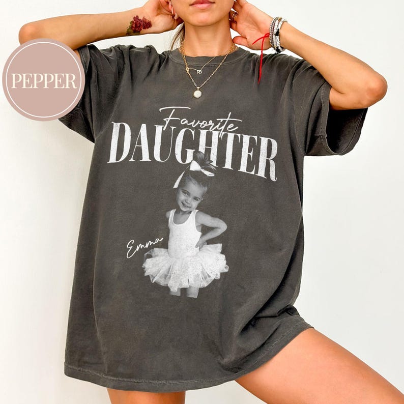 May include: Dark gray t-shirt with the words "Favorite Daughter" in white block letters. Below the text is a black and white image of a young girl in a tutu. The girl's name, "Emma," is written below the image.
