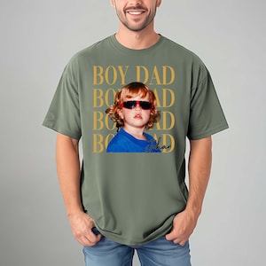 Pode incluir: Camiseta verde-oliva com um gr&aacute;fico de uma crian&ccedil;a usando &oacute;culos de sol. O texto "BOY DAD" &eacute; impresso em dourado acima da imagem da crian&ccedil;a, com o nome "Johan" em it&aacute;lico abaixo.
