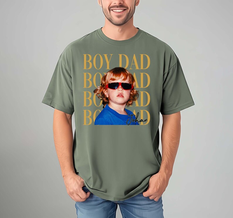 Pode incluir: Camiseta verde-oliva com um gr&aacute;fico de uma crian&ccedil;a usando &oacute;culos de sol. O texto "BOY DAD" &eacute; impresso em dourado acima da imagem da crian&ccedil;a, com o nome "Johan" em it&aacute;lico abaixo.