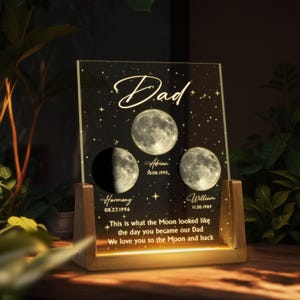 May include: A personalized acrylic night light with a wooden base. The design features three moon phases, names, and dates, with the word "Dad" at the top. The text reads "This is what the Moon looked like the day you became our Dad."