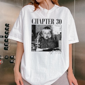 May include: White t-shirt with a black and white photo of a young child with pigtails sticking out their tongue. The text "CHAPTER 30" is above the photo, and "est. 1996" is below it. The shirt is a casual, oversized fit.