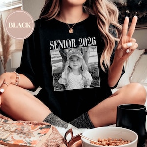 May include: Black t-shirt featuring the text "SENIOR 2026" above a black and white photograph of a young girl. The word "BLACK" is in a circle in the upper left. The shirt is a relaxed fit.