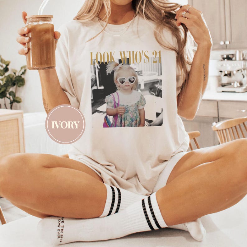 May include: Ivory-colored t-shirt with the text "LOOK WHO'S 21" in gold lettering. The shirt features a black and white image of a young girl wearing heart-shaped sunglasses. The person in the image is holding a glass of iced coffee.