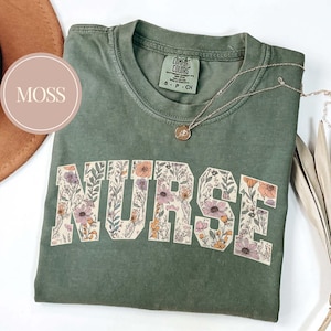 May include: A moss green t-shirt with the word "NURSE" in large, floral-filled letters. The shirt has a crew neck and short sleeves. A delicate silver necklace with a small pendant rests on the shirt. The shirt is folded and placed on a white surface.