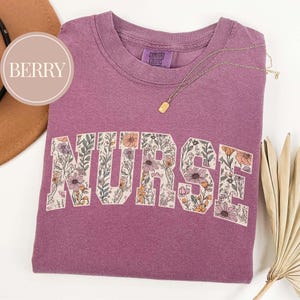 May include: A berry-colored t-shirt with the word "NURSE" in a floral design. The shirt has a crew neck and a small tag that says "COMFORT COLORS". A gold necklace with a rectangular pendant is draped across the shirt.